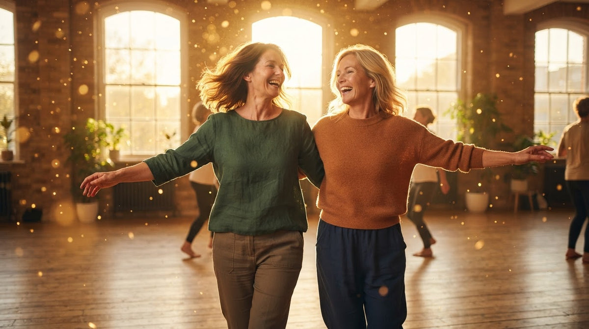 Zwei Frauen um die 50 tanzen an einem Frühlingsnachmittag lachend nebeneinander in einem hellen Tanzstudio