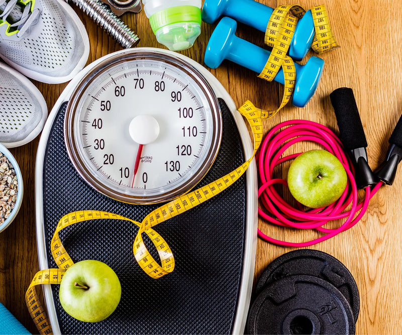 Foto einer Waage, umgeben von weiteren kleinen Fitnessgeräten und zwei Äpfeln
